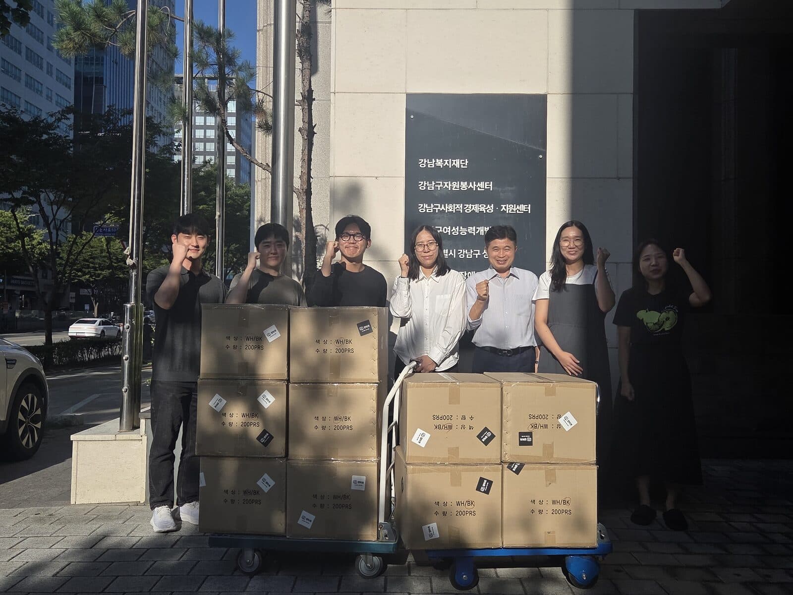 강남복지재단 건물 앞에서 일곱 사람이 양말 상자를 쌓은 운반대 두 대를 사이에 두고 서 있다. 상자마다 '수량 : 200PRS' 라벨이 붙어 있다.