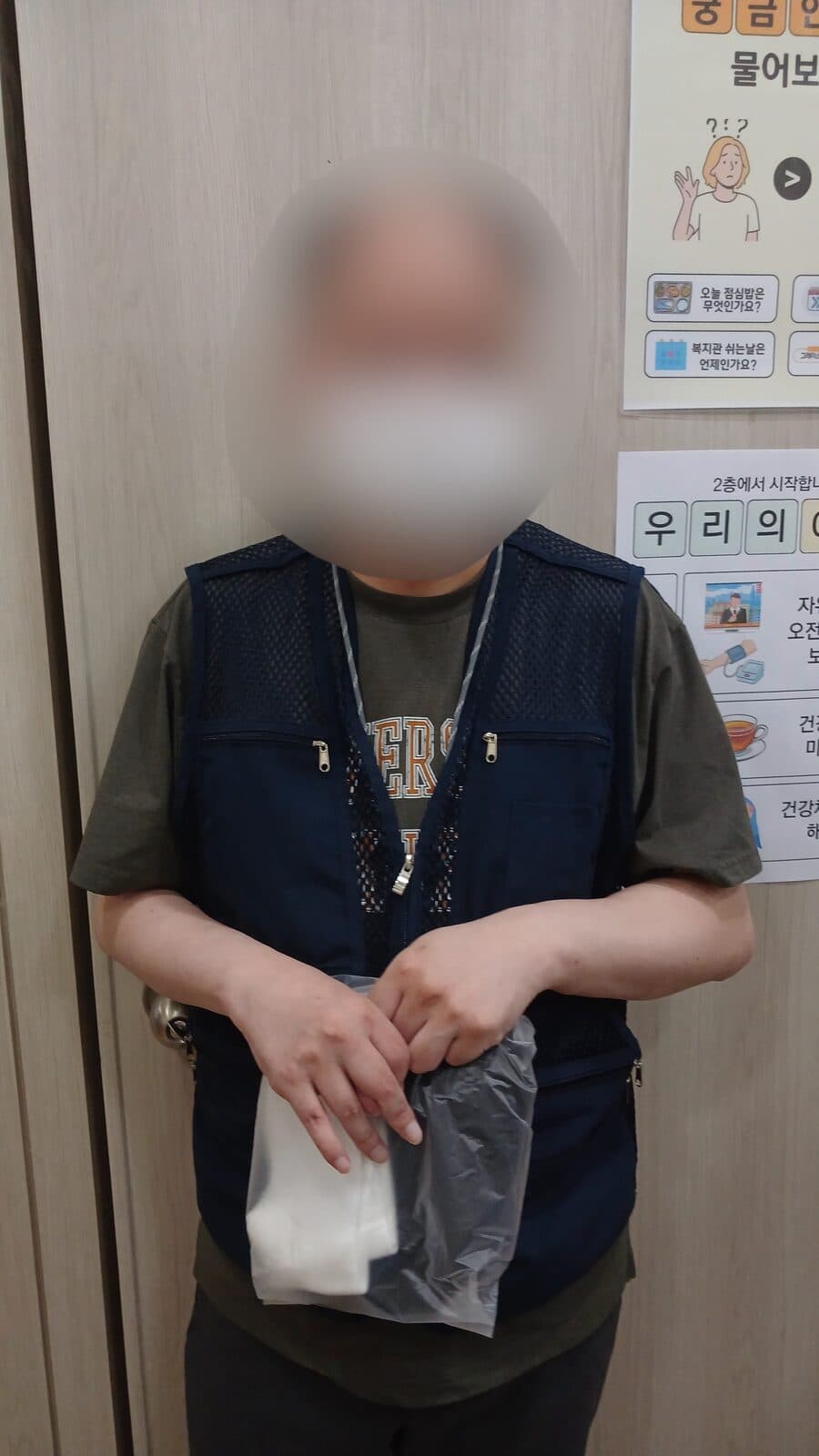 복지관 실내에서 조끼를 입은 사람이 양말이 담긴 봉투를 두 손으로 들고 있다.
